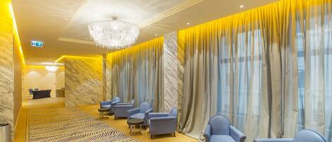 Un vestíbulo de hotel con un mostrador de recepción de mármol, sillones azules, una alfombra estampada y un candelabro.