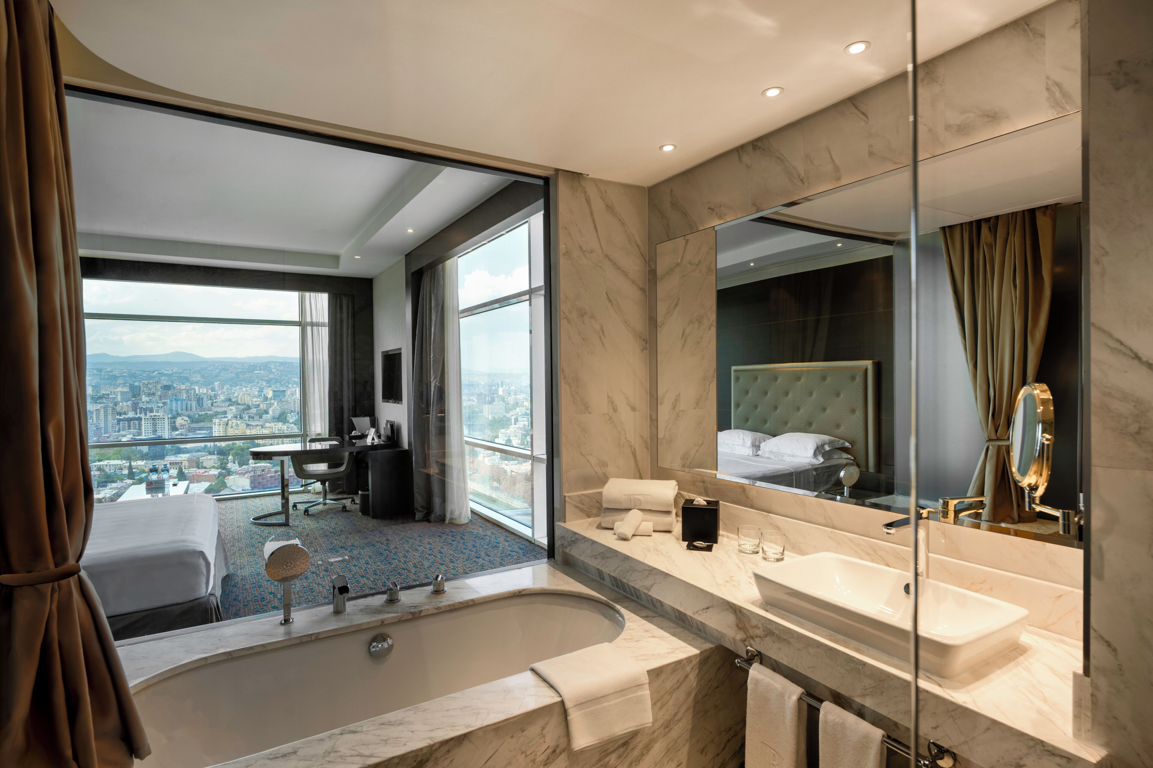A modern hotel bathroom with a large bathtub, a vanity with a mirror, and a view of the city through large windows.