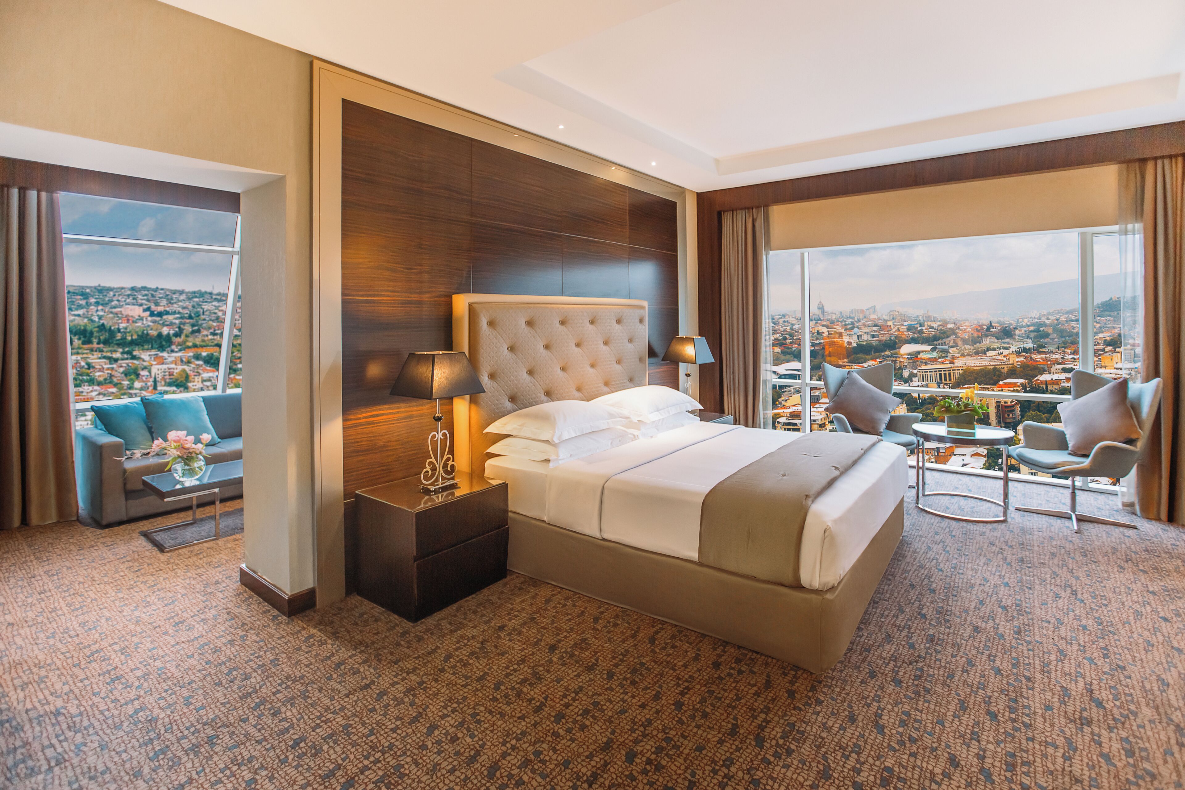 A hotel room with a large bed, a sofa, a small table, and a view of a cityscape.
