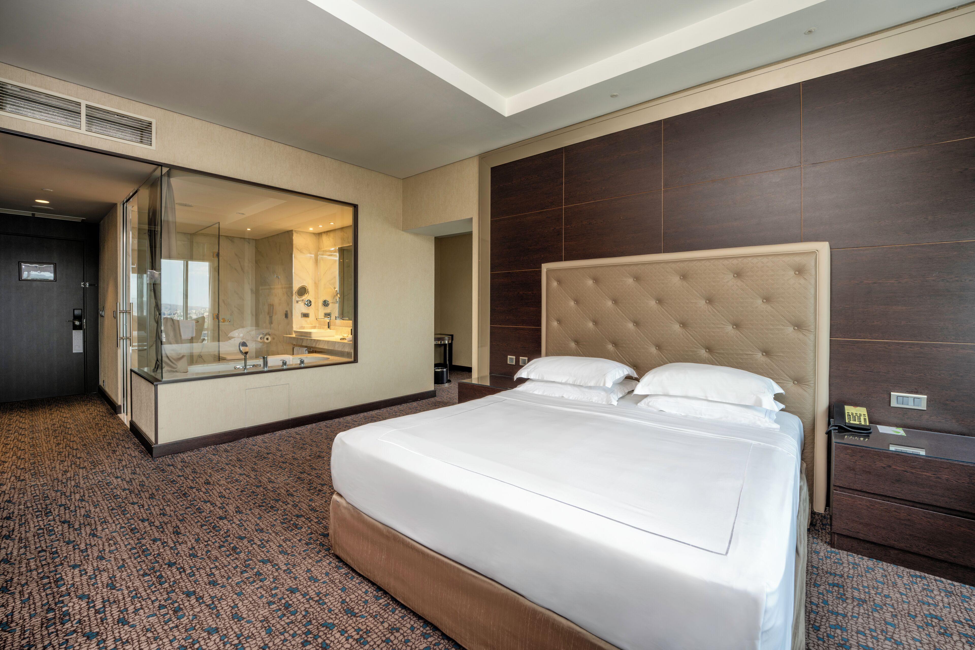 A hotel room with a large bed, a glass-enclosed shower, a dark wood headboard, and a minibar.