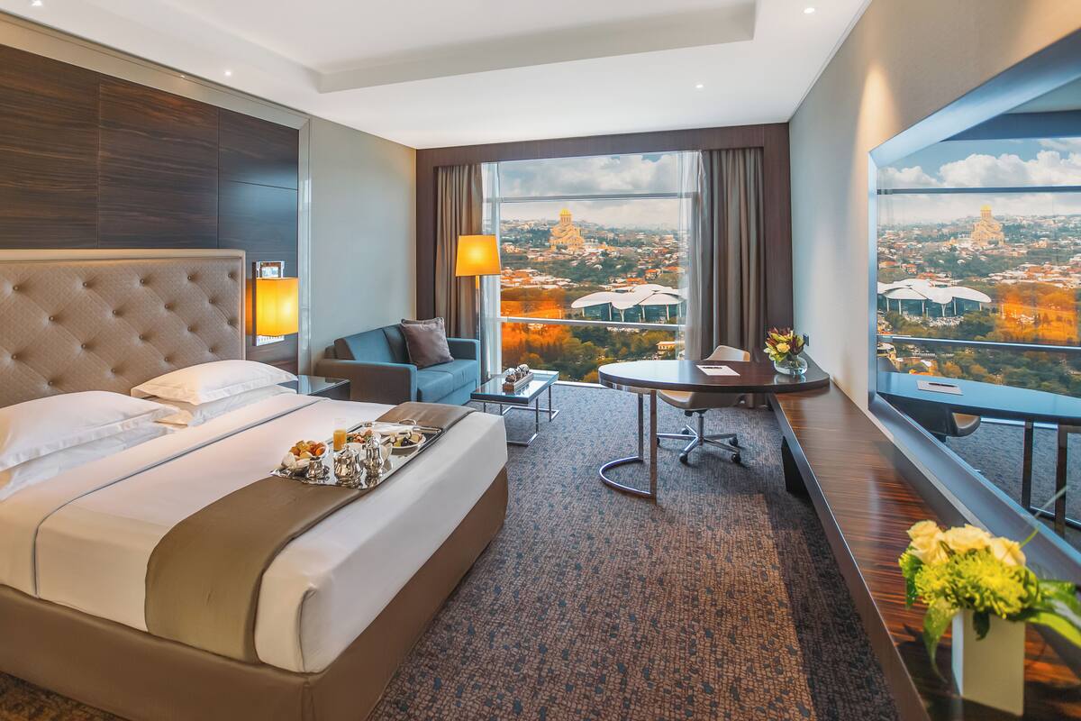 Ein Hotelzimmer mit einem großen Bett, einer Sitzecke, einem Esstisch und Blick auf eine Stadtlandschaft.