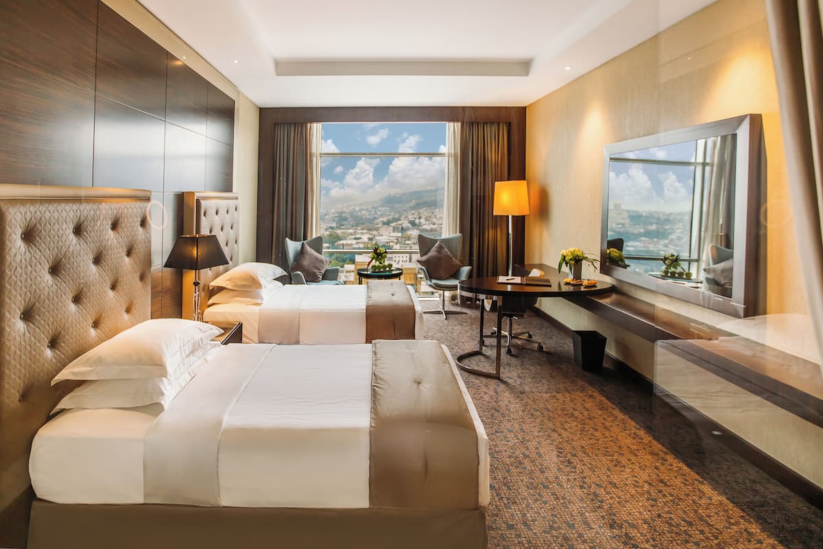 Ein Hotelzimmer mit zwei Betten, einem Schreibtisch, einem Stuhl, einer Lampe und einem großen Fenster mit Blick auf die Stadt.