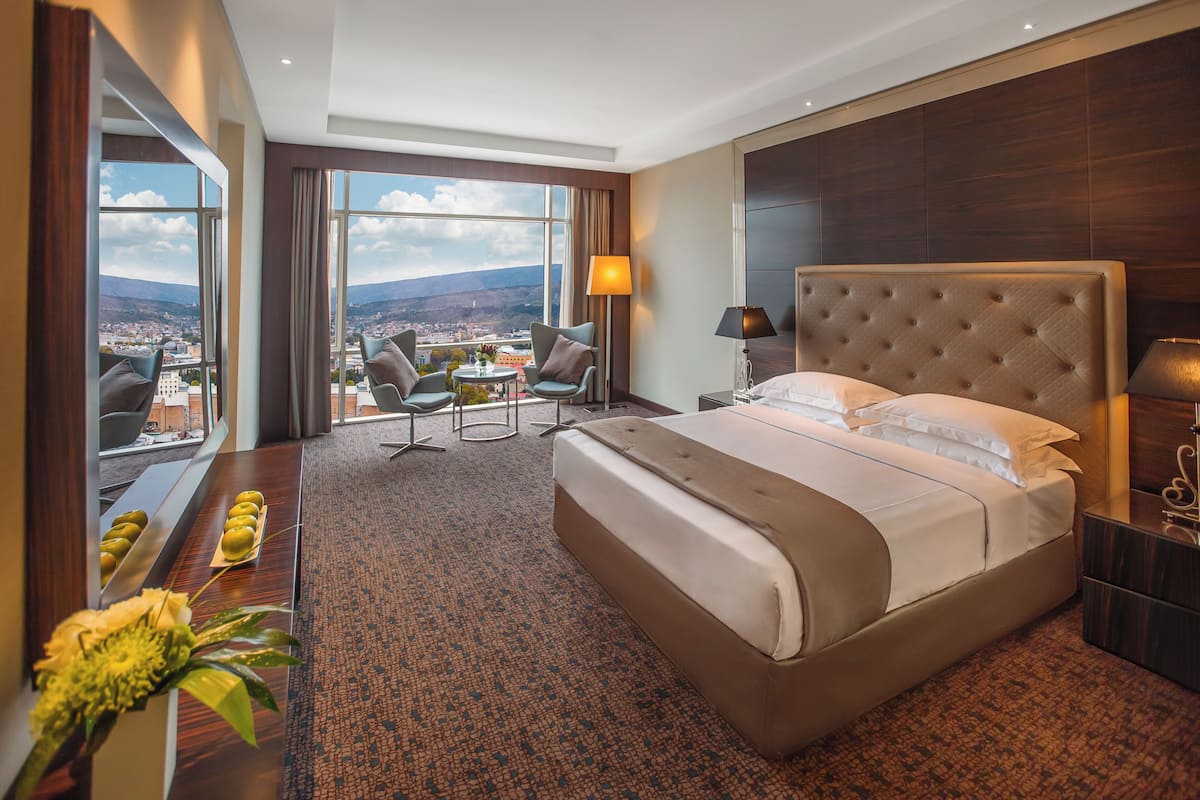 Ein Hotelzimmer mit einem großen Bett, einem Schreibtisch mit Stuhl, einem kleinen Tisch mit Stühlen und Blick auf die Stadt durch große Fenster.
