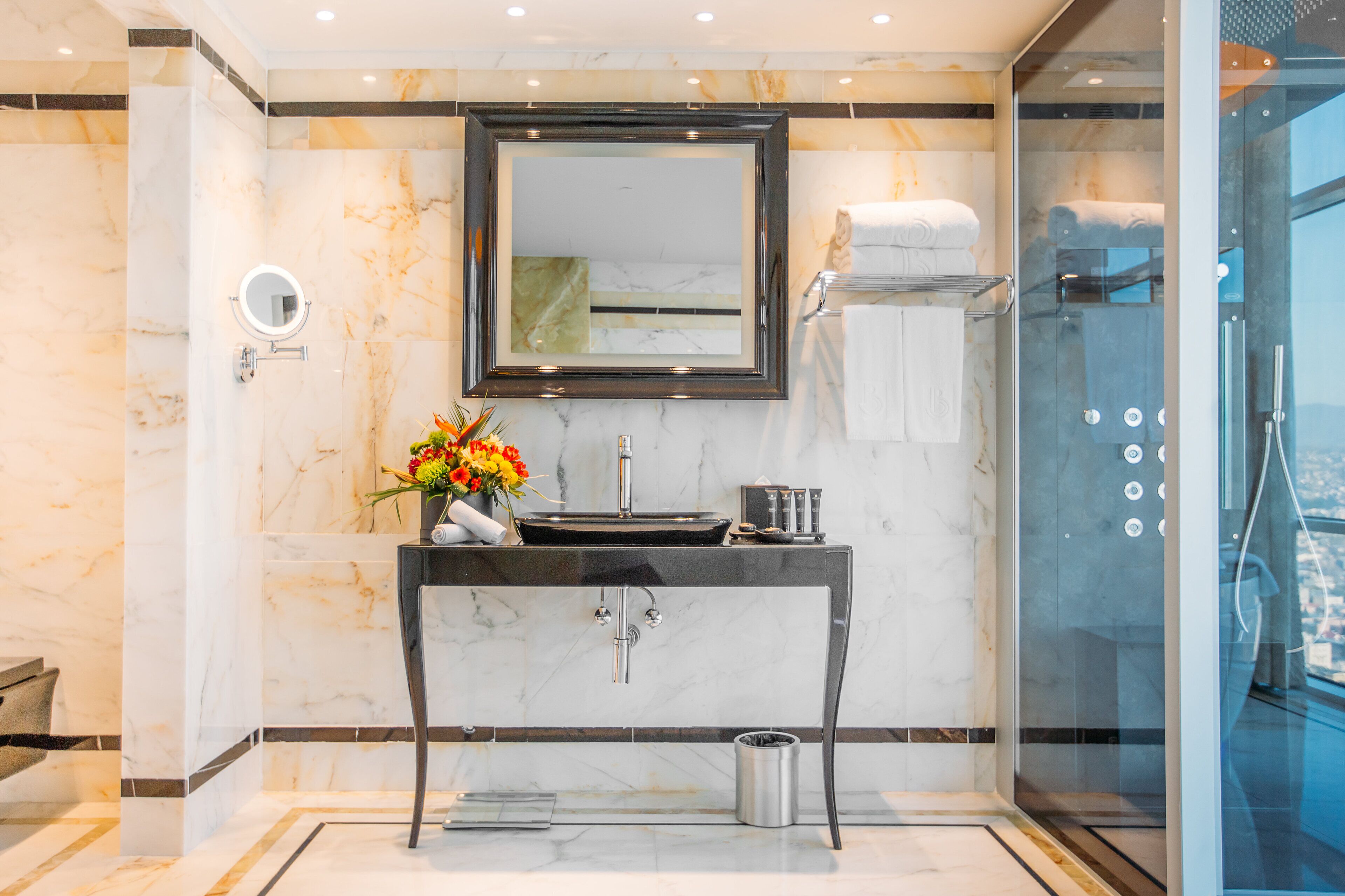 A modern bathroom with a marble wall, a large mirror, a sink with a vase of flowers, and a glass-enclosed shower.