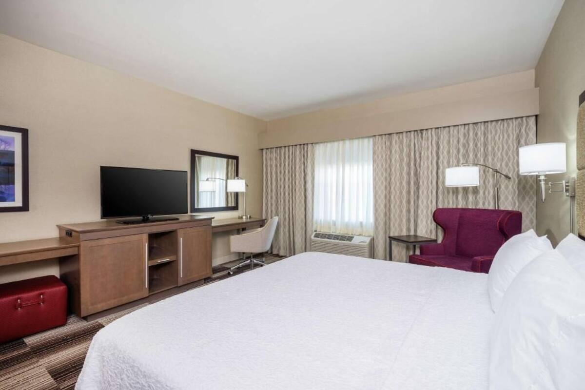 A hotel room with a bed, a desk with a television, a chair, a red armchair, and a small table with a lamp.