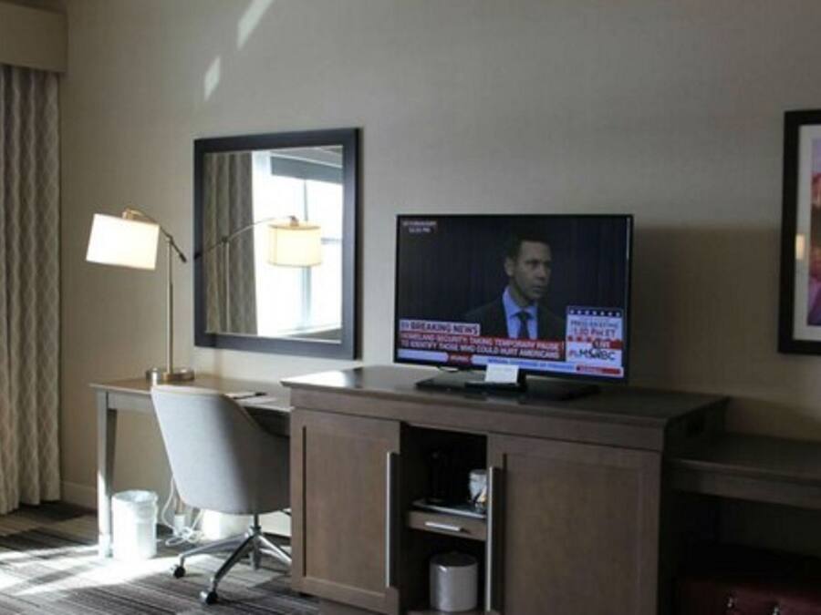 A hotel room with a television on a wooden stand, a desk with a chair, a lamp, and a framed picture on the wall.