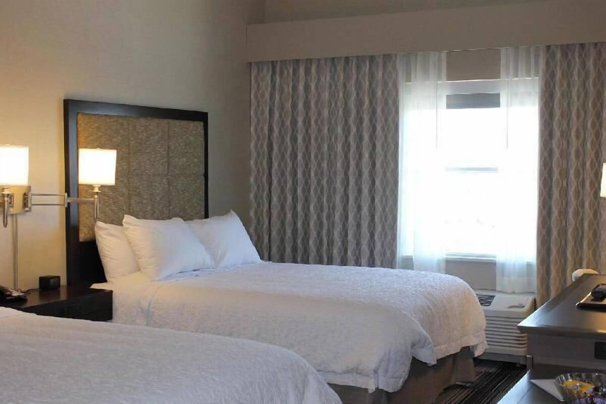 A hotel room with two beds, a desk, a lamp, and a large window with curtains.