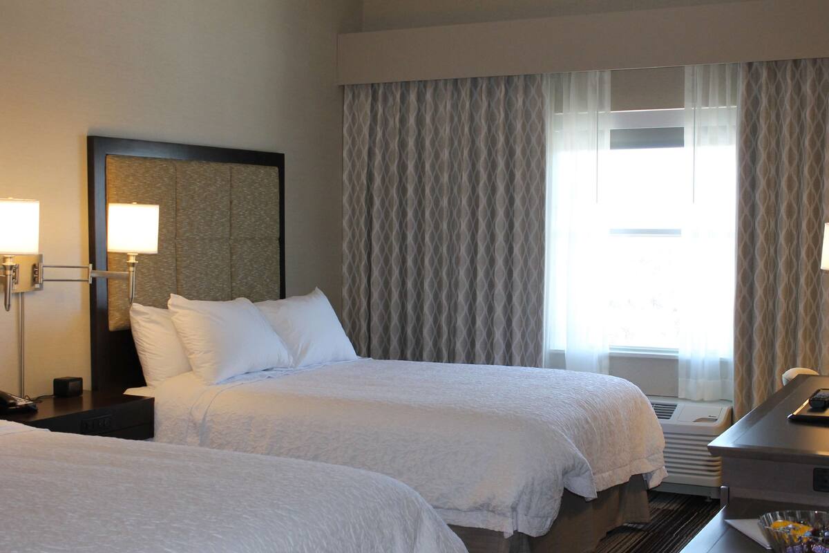 A hotel room with two beds, a desk, a lamp, and a large window with curtains.