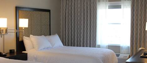 Habitación de hotel con dos camas, un escritorio, una lámpara y un ventanal grande con cortinas.