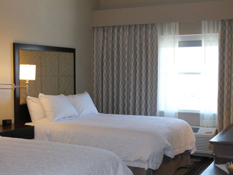Ein Hotelzimmer mit zwei Betten, einem Schreibtisch, einer Lampe und einem großen Fenster mit Vorhängen.