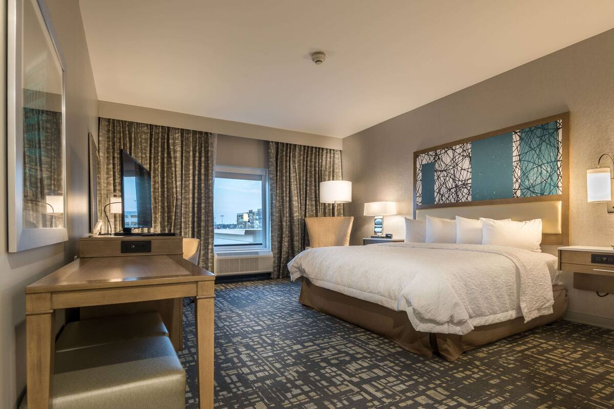 Une chambre d’hôtel avec un grand lit, un bureau, une chaise, une fenêtre avec des rideaux et une vue sur les bâtiments environnants.