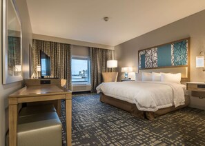 A hotel room with a large bed, a desk, a chair, a window with curtains, and a view of buildings outside.
