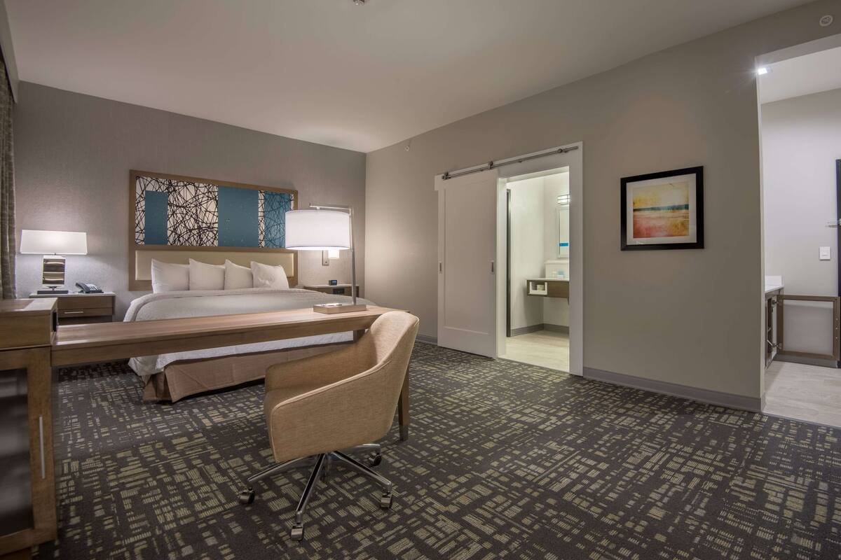 Une chambre d’hôtel avec un lit, un bureau, une chaise, une lampe, un tableau et une salle de bain visible à travers une porte ouverte.