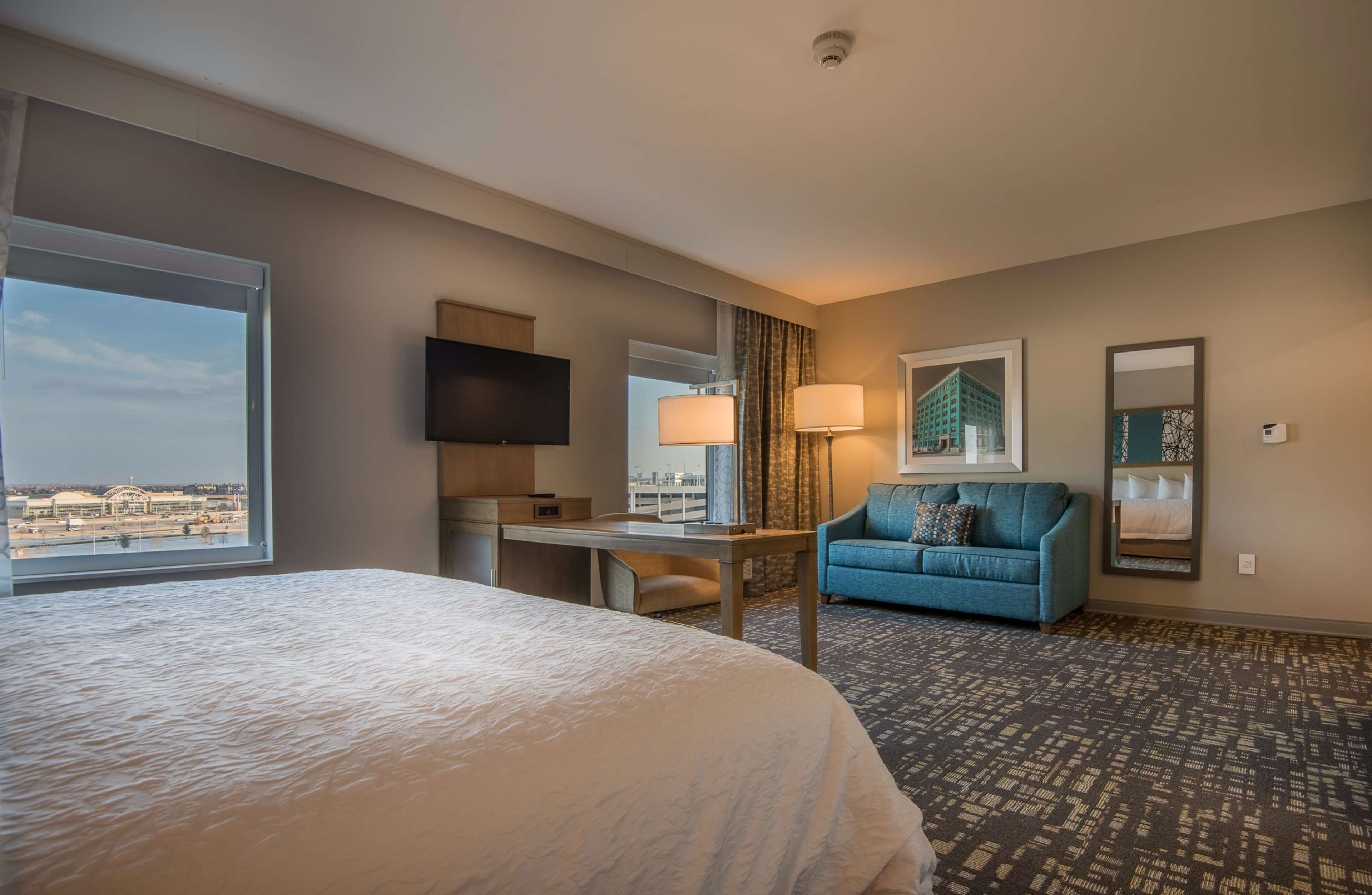 A hotel room with a large bed, a desk, a blue sofa, a TV, and a view of the city.