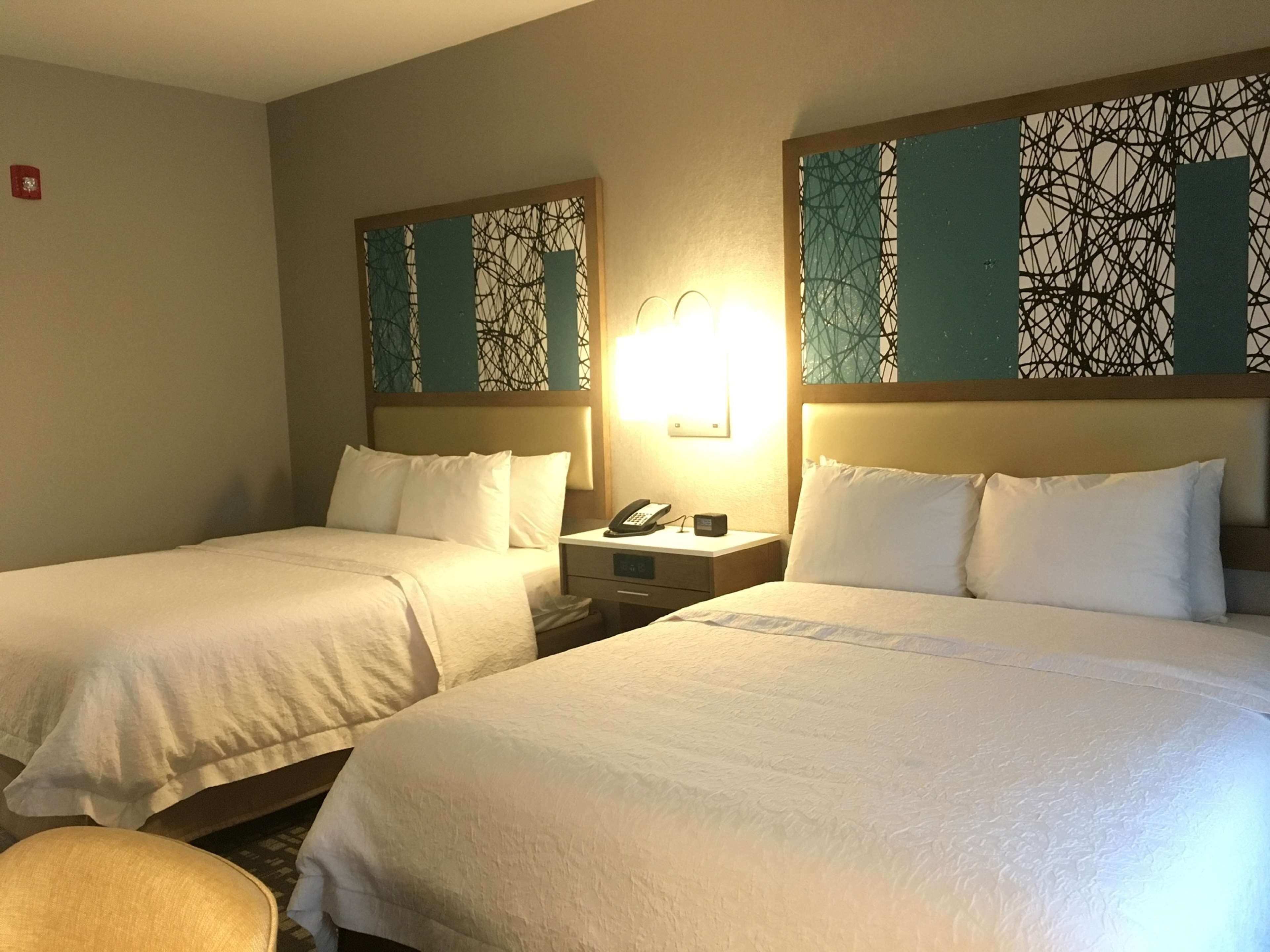 A hotel room with two beds, a nightstand with a phone, and a wall-mounted light.