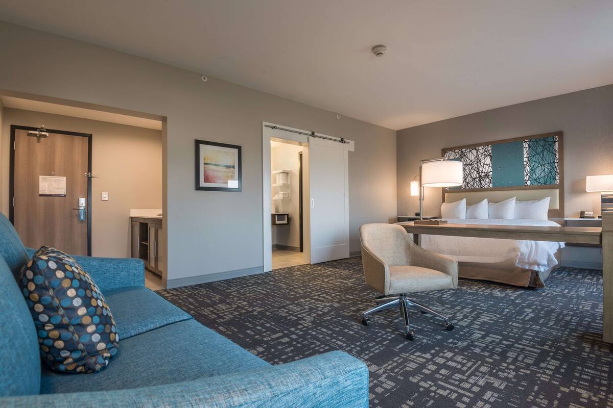 Une chambre d’hôtel avec un canapé bleu, un bureau avec une lampe, un lit et une porte donnant sur une salle de bain.