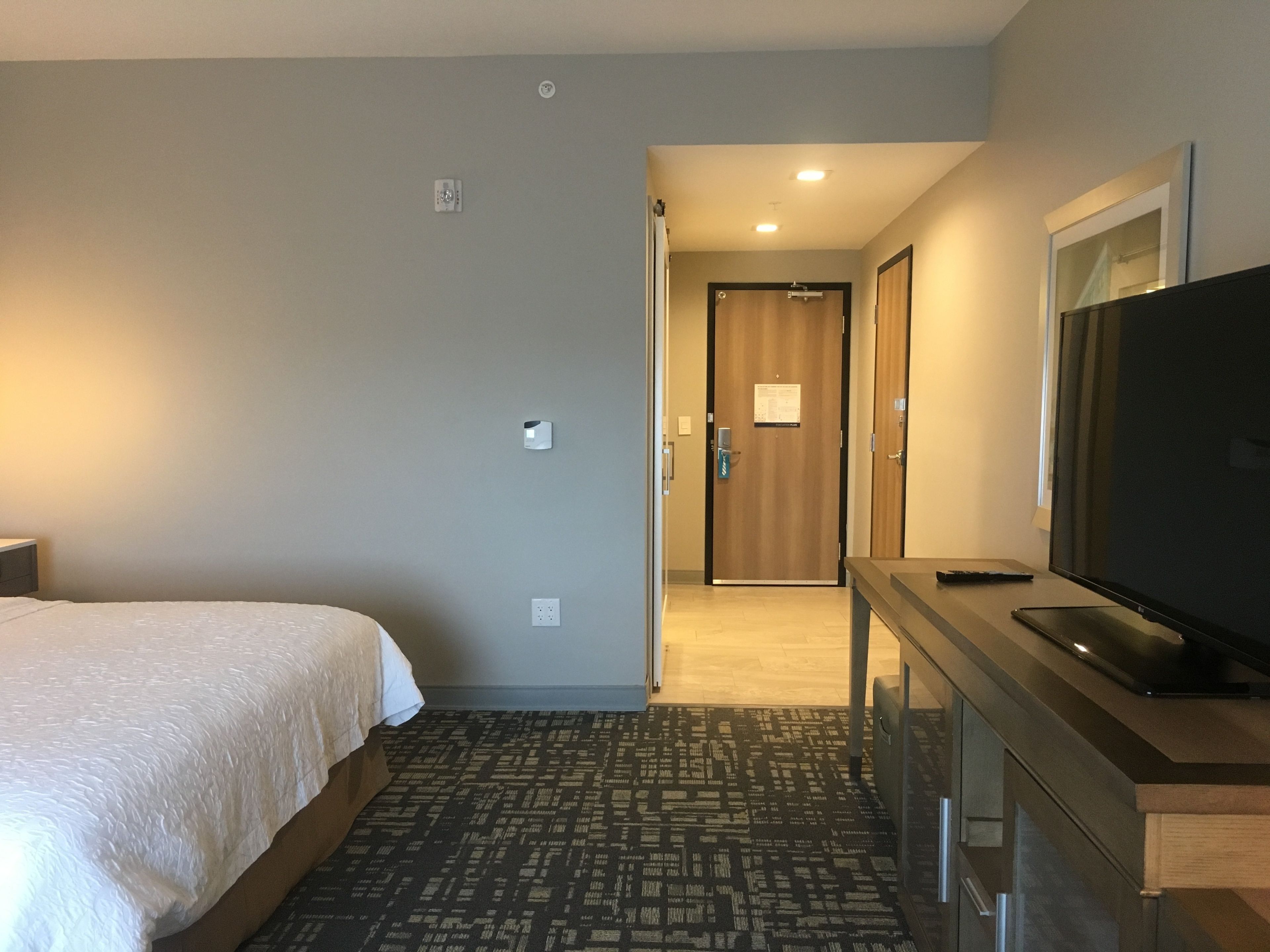 Habitación de hotel con cama, televisión, escritorio y puerta que da a un pasillo.