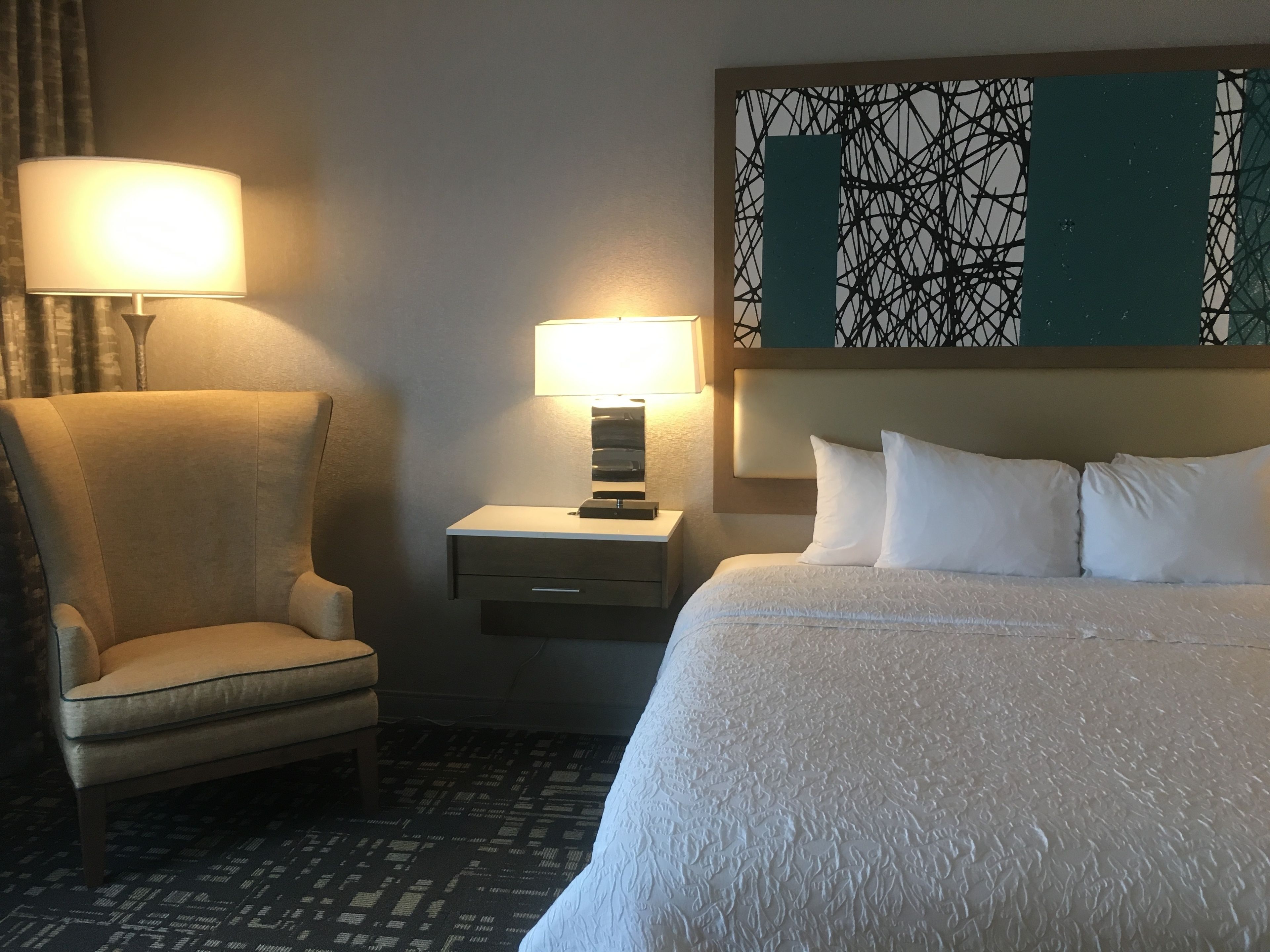 Quarto de hotel com cama, poltrona, mesa de cabeceira com abajur e uma obra de arte na parede.