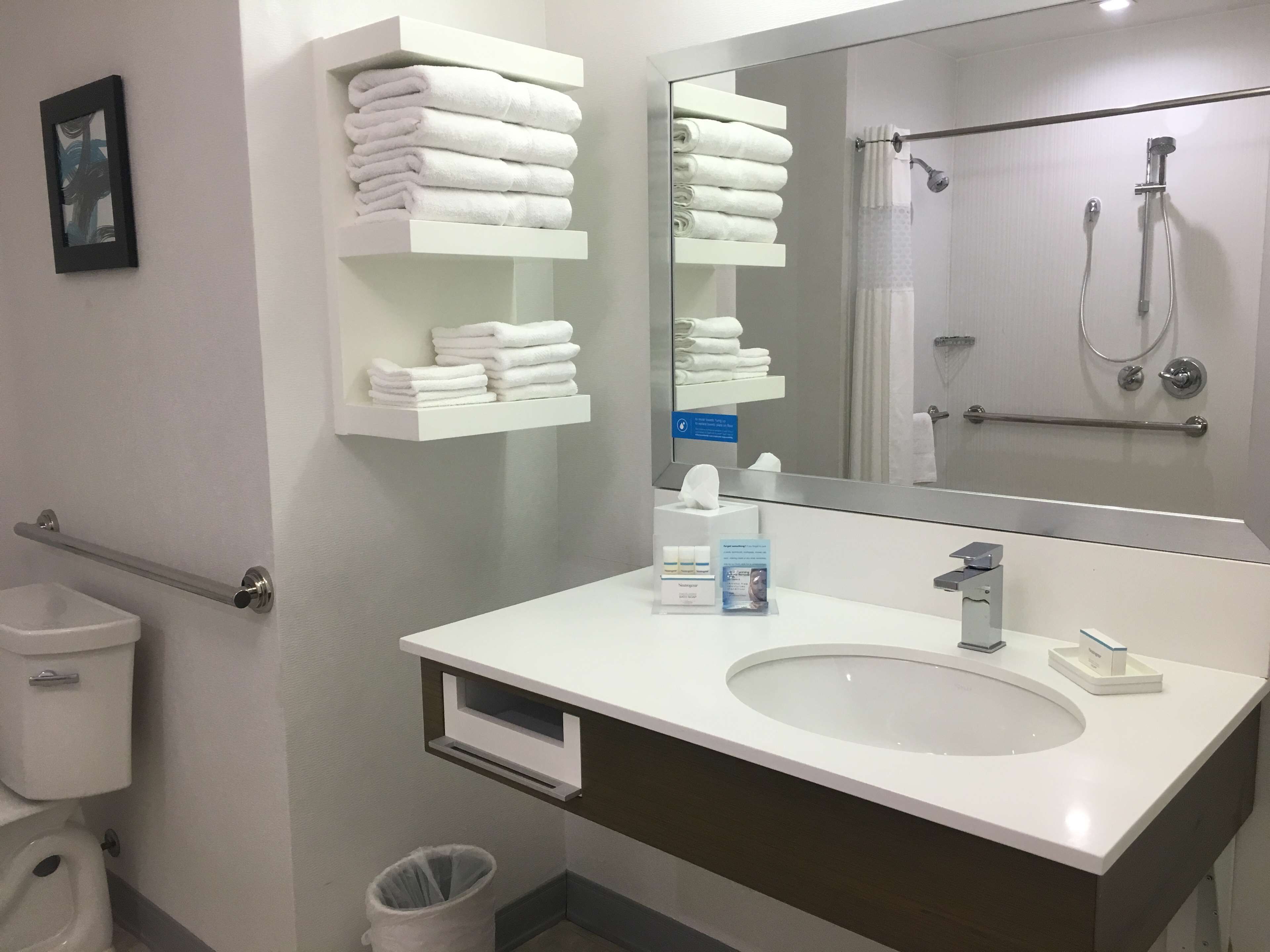 Une salle de bain moderne avec un lavabo blanc, un grand miroir et une douche. On peut voir des serviettes blanches pliées sur une étagère, et un sèche-mains est installé.