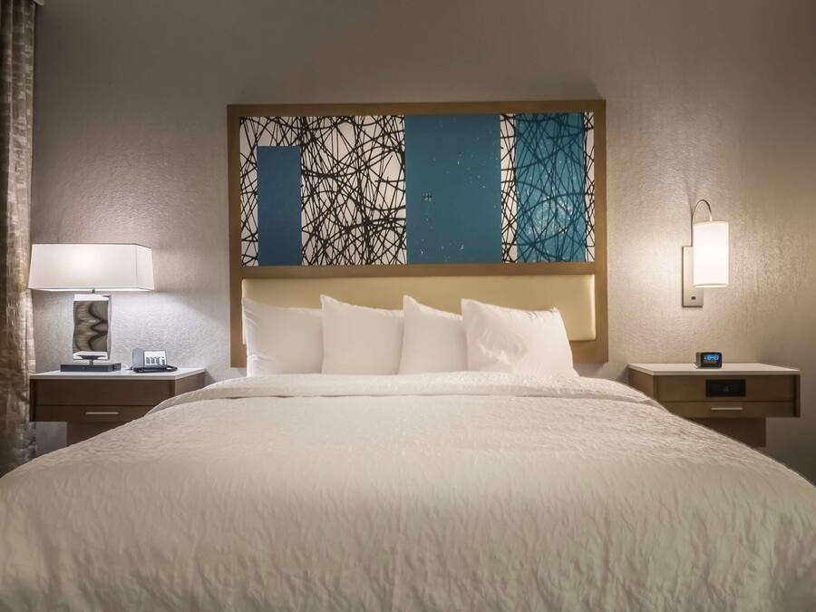 Habitación de hotel con una cama grande, una mesita de noche con lámpara, un reloj de pared y un cabecero decorativo.