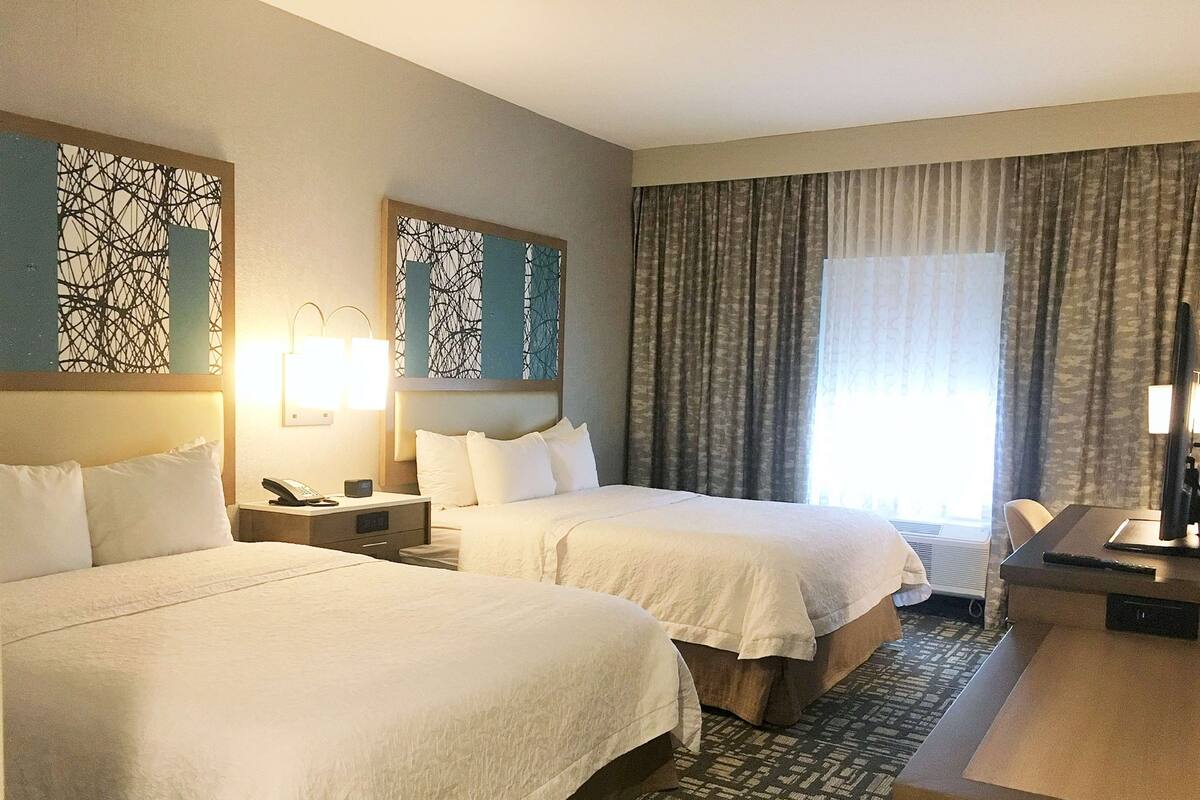 Une chambre d’hôtel avec deux lits, un bureau, une chaise, une lampe et une fenêtre avec des rideaux.