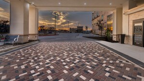 A spacious outdoor area with a patterned brick floor, a covered walkway, and a view of the sunset over a parking lot and buildings. - Hampton Inn & Suites Dallas-The Colony, TX (The Colony)