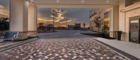 Un espacio exterior amplio con piso de ladrillo estampado, un pasillo cubierto y vista al atardecer sobre un estacionamiento y edificios.