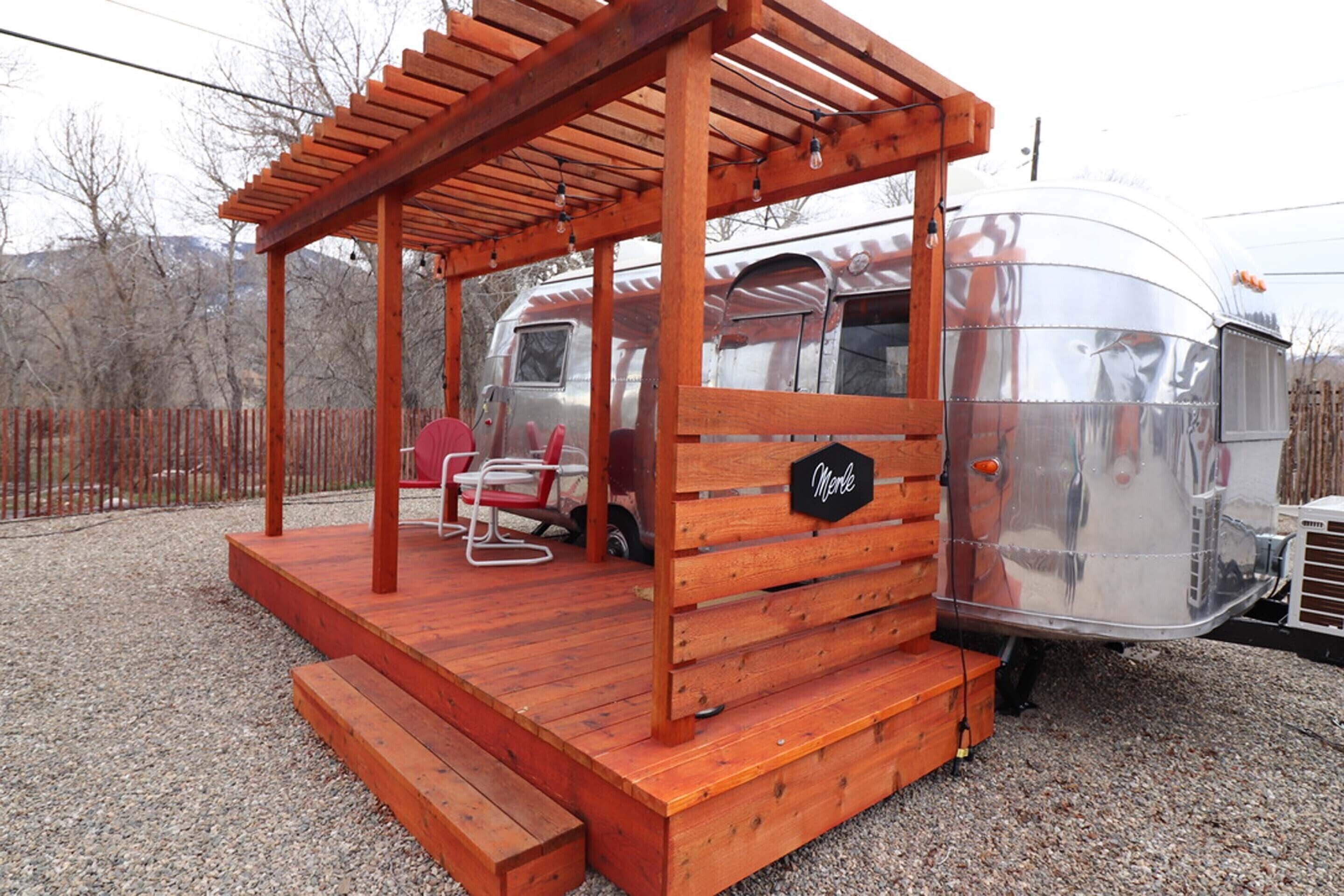 A wooden structure with a bench and a sign labeled "Merle" in front of a silver trailer.