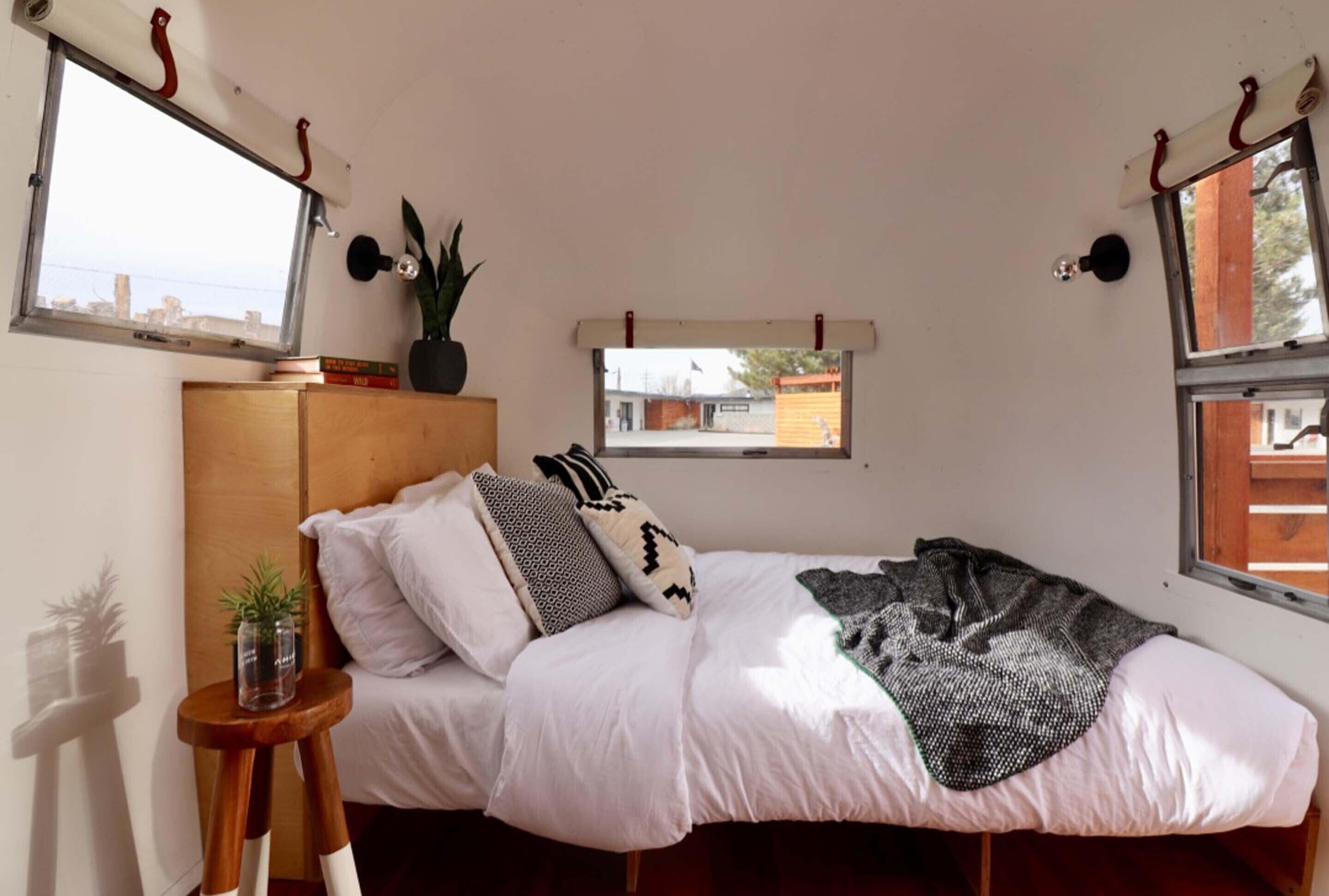 Un dormitorio con una cama, una mesita de noche de madera, una planta en maceta y un ventanal con vista a un edificio.
