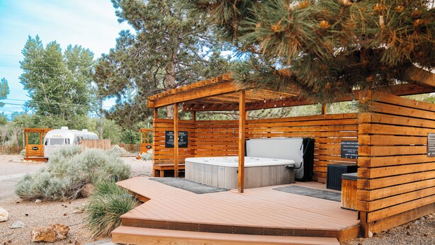 A wooden structure with a hot tub and a covered seating area, surrounded by trees and a gravel path. - The Amigo Motor Lodge (Salida)