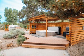 A wooden structure with a hot tub and a covered seating area, surrounded by trees and a gravel path. - The Amigo Motor Lodge (Salida)