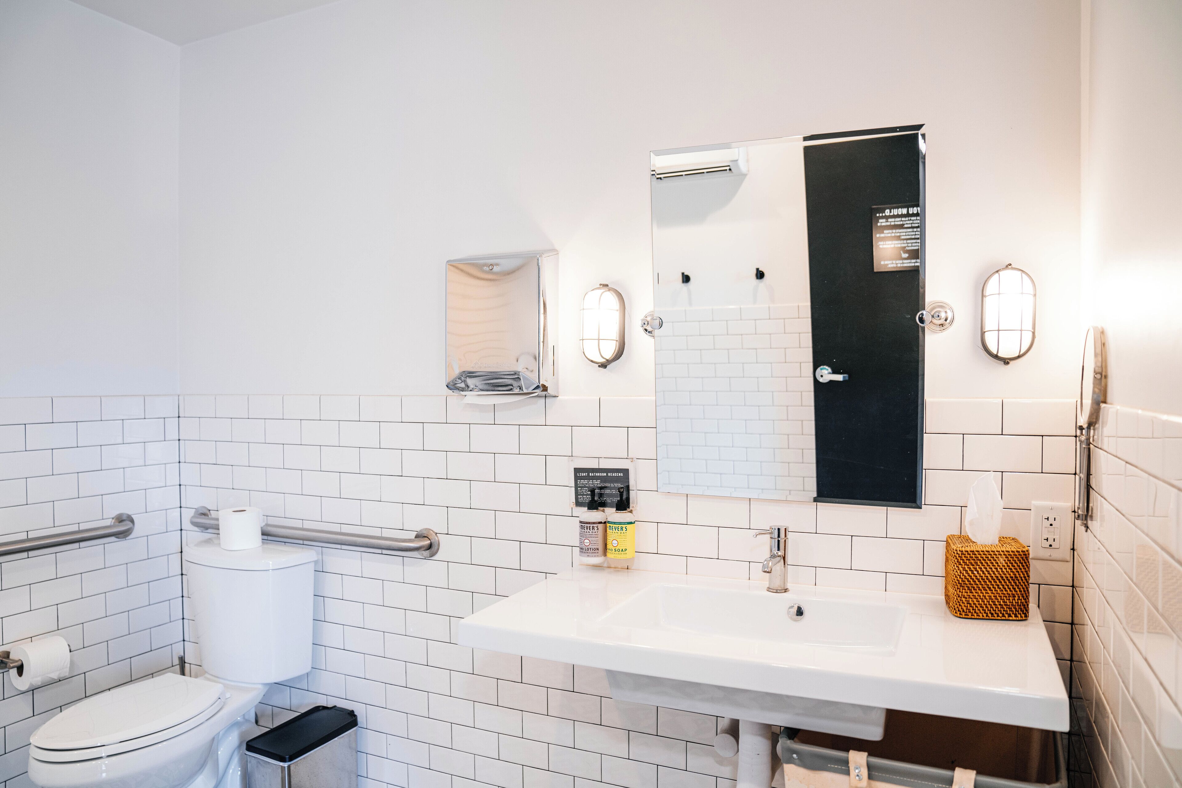 Un baño moderno con pared de azulejos blancos, un lavamanos blanco, un espejo, un inodoro y un pasamanos.