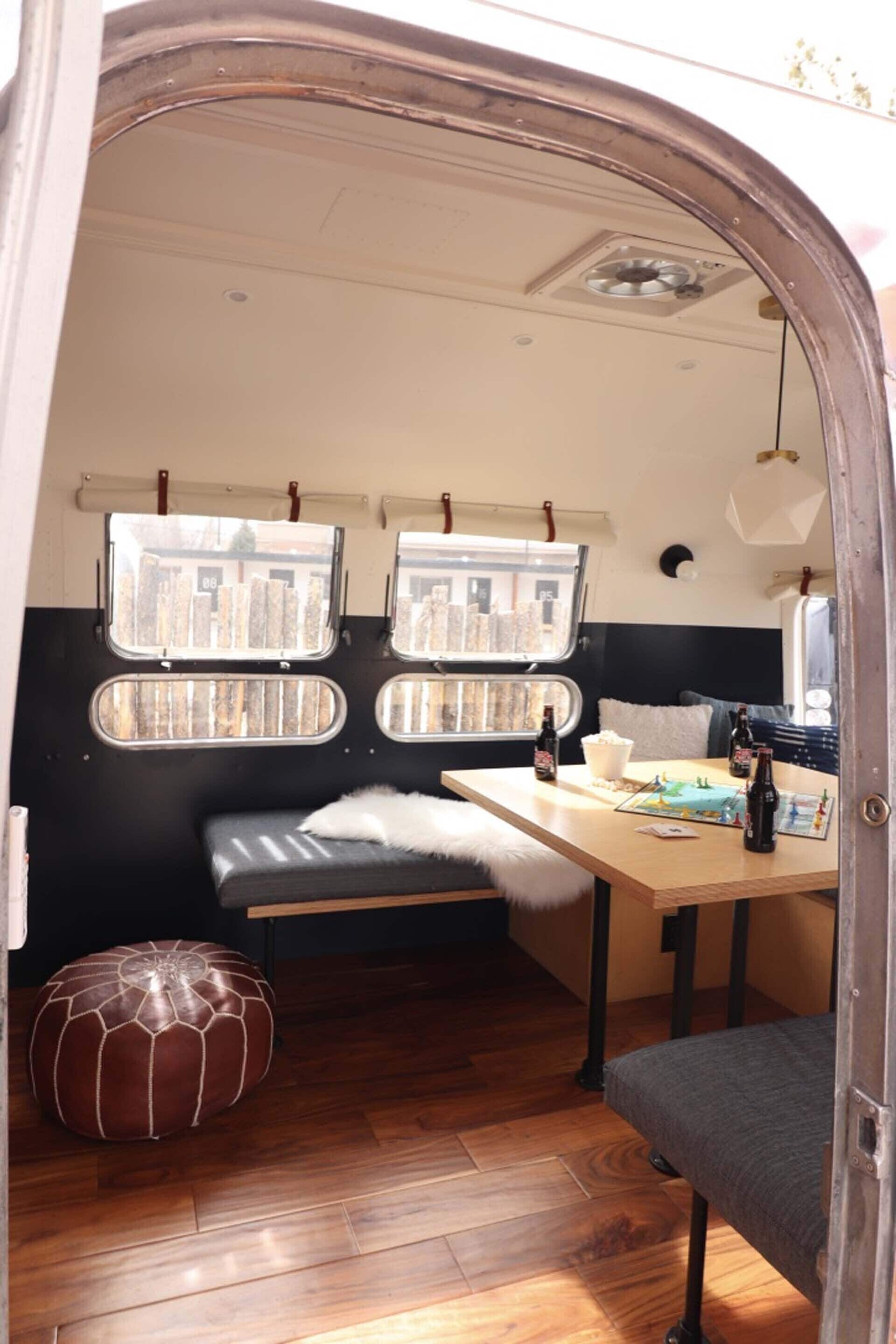 A cozy interior with a wooden floor, a bench, a table with bottles, and two windows with views of buildings.