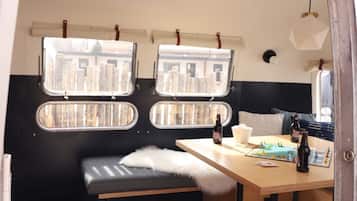A cozy interior with a wooden floor, a bench, a table with bottles, and two windows with views of buildings.