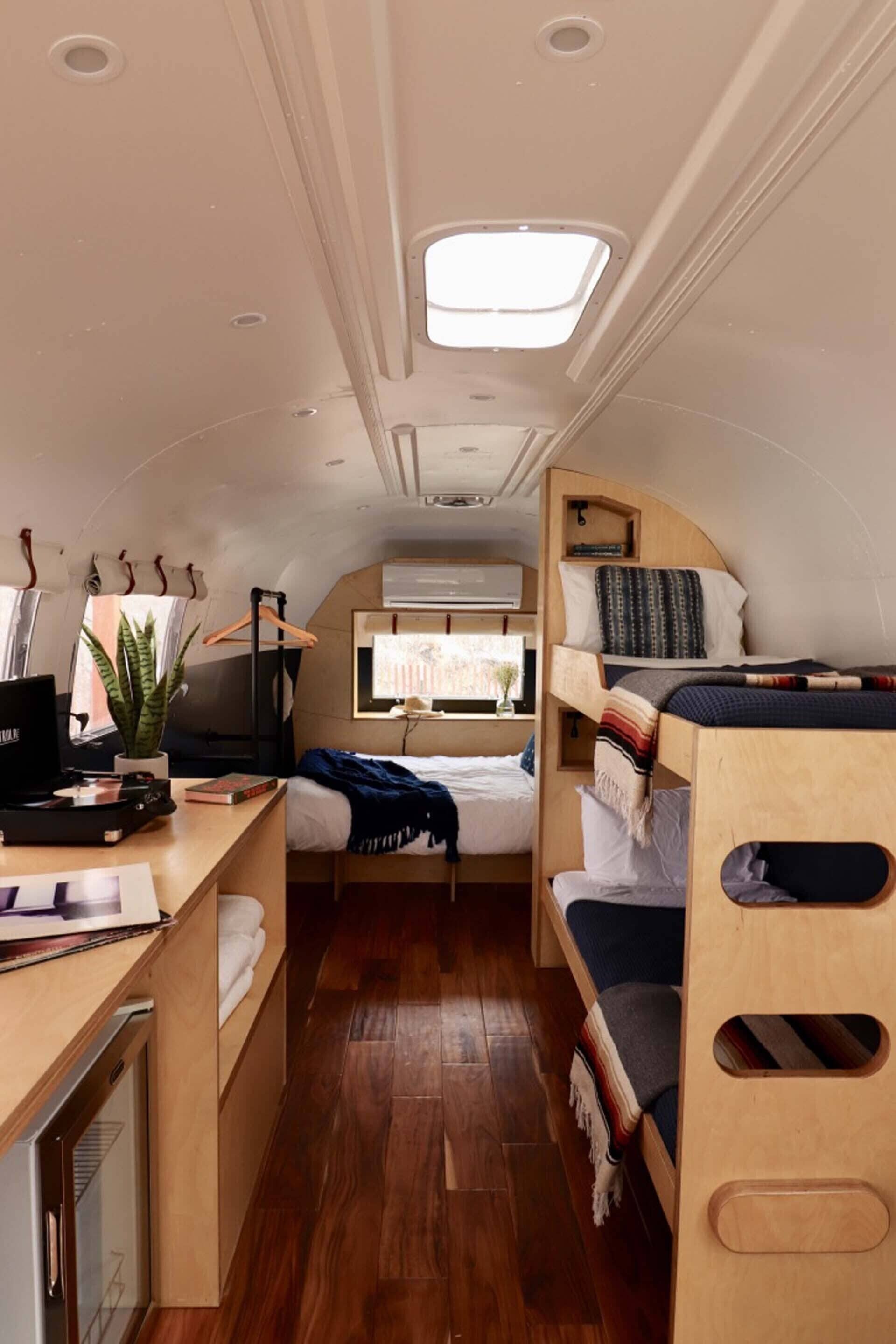 A compact RV interior with a kitchenette, a bed, a desk with a computer, and a window providing natural light.
