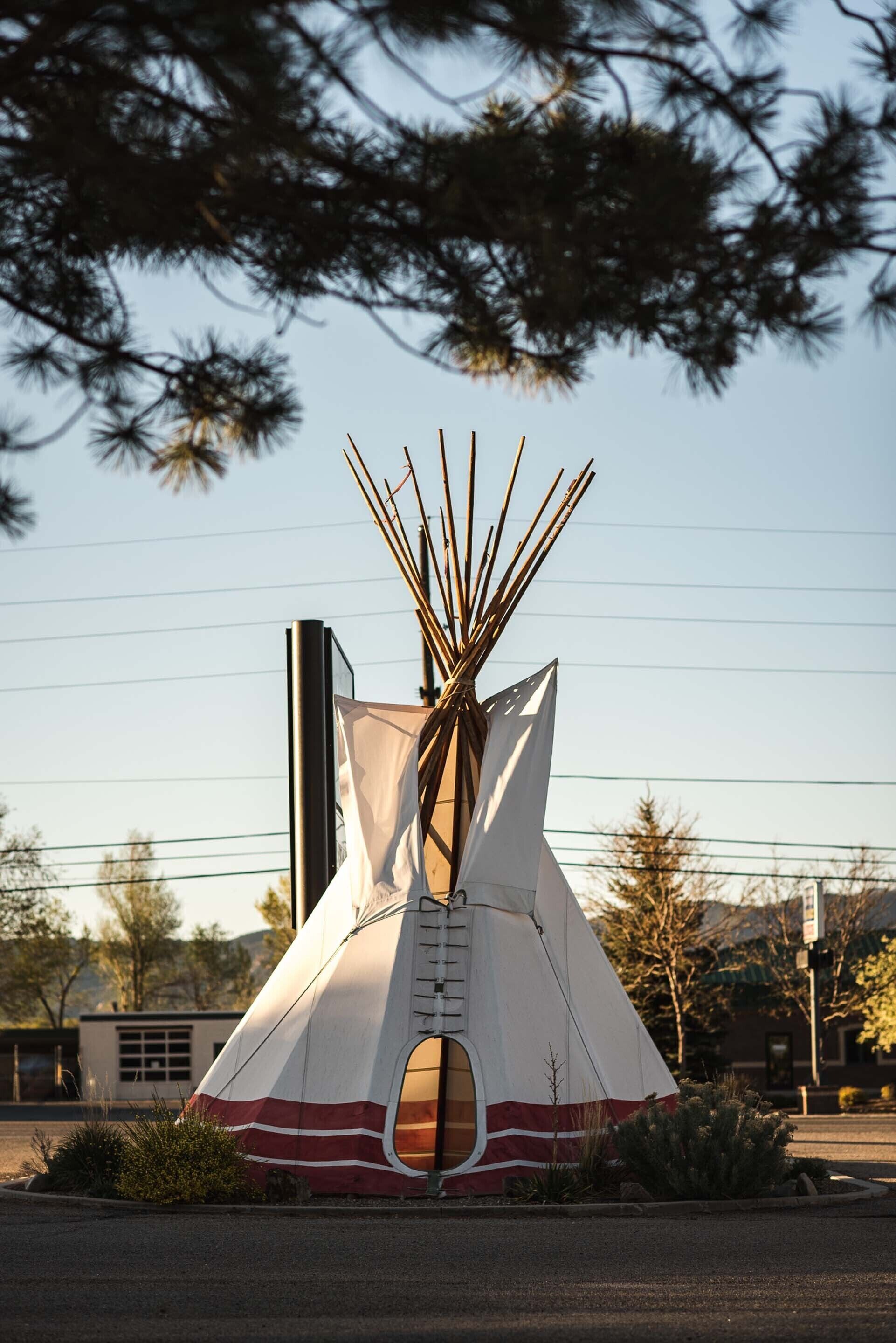 A tipi-style tent with a ladder, set in a landscaped area with trees and a building in the background.