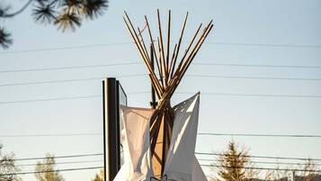 A tipi-style tent with a ladder, set in a landscaped area with trees and a building in the background.