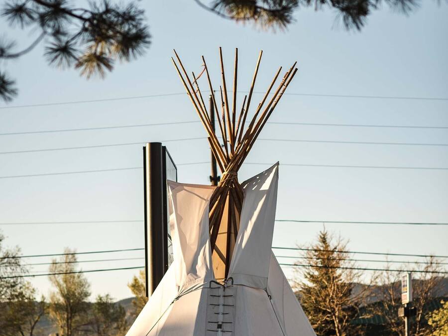 A tipi-style tent with a ladder, set in a landscaped area with trees and a building in the background.