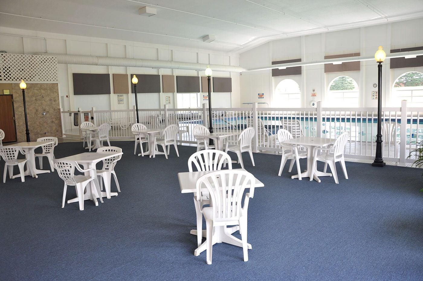 A spacious indoor area with white plastic chairs and tables arranged in rows, a pool visible through a white fence, and a blue carpeted floor.