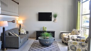 A living room with a sofa, armchair, and a round coffee table with a plant. There is a flat-screen TV on the wall and a window with a curtain. - Dunes Express Inn & Suites (Hart)
