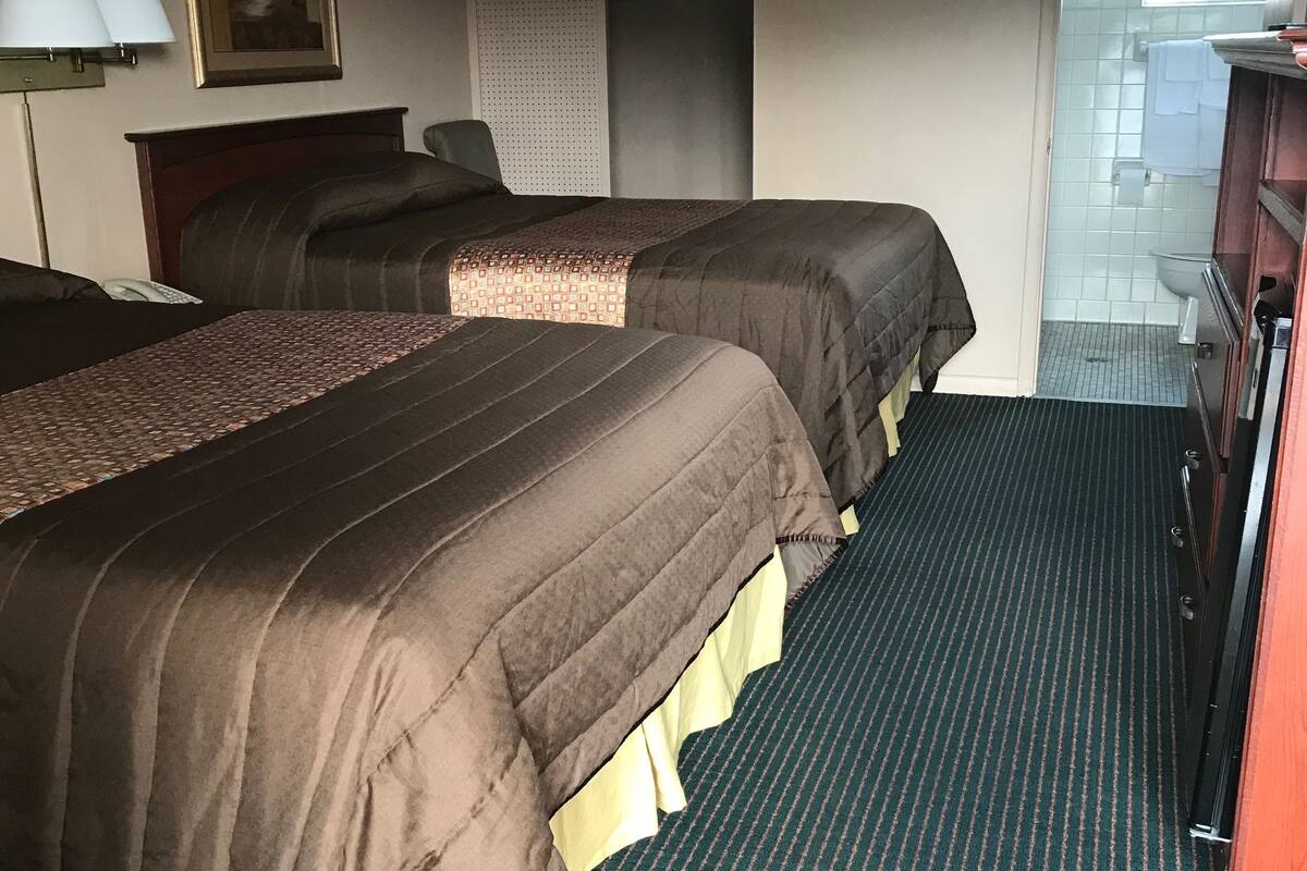 A hotel room with two beds, a desk, a chair, a painting on the wall, and a bathroom visible through an open door.