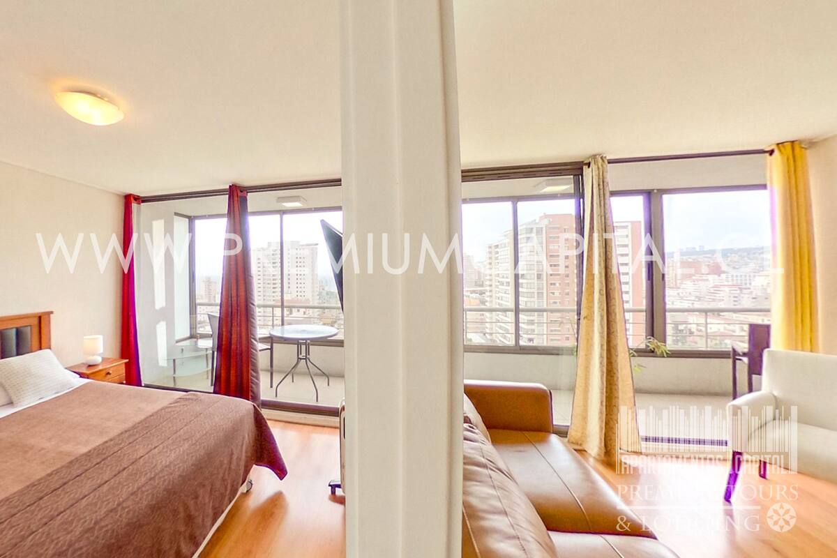 A hotel room with a bed, a sofa, a dining table, and large windows offering a city view.