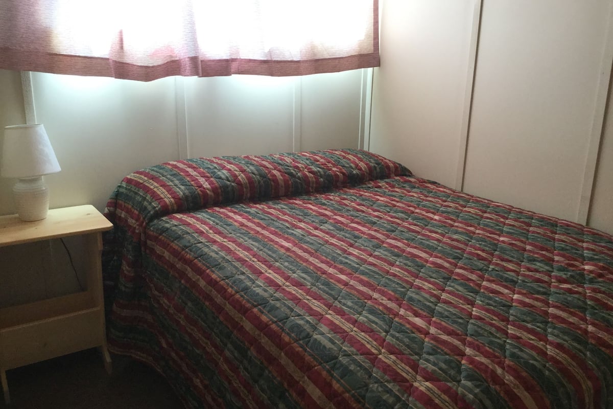 Una habitación pequeña con una cama individual, colcha a rayas, mesita de noche con lámpara y ventana con cortinas traslúcidas.