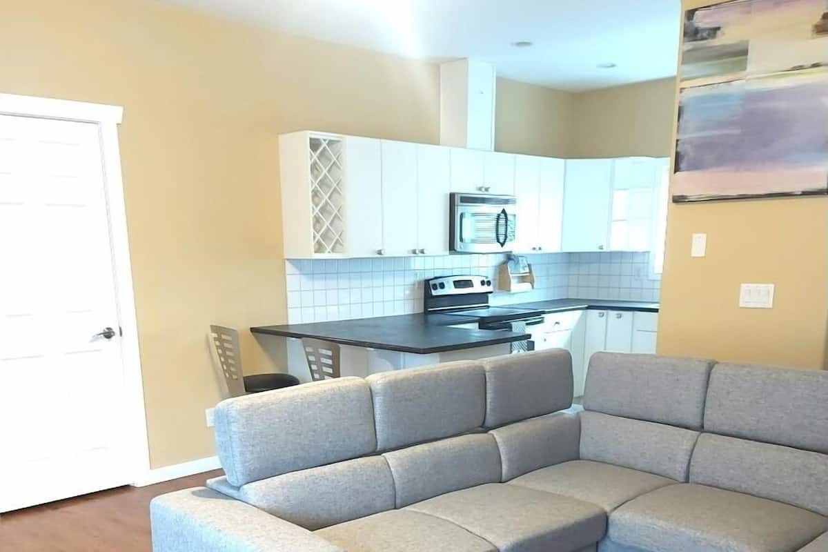 Una sala de estar con un sofá seccional gris, piso de madera, una cocina con armarios blancos y una puerta.