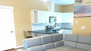 A living room with a grey sectional sofa, wooden flooring, a kitchen with white cabinets, and a door. - Cedars Motel (Princeton)