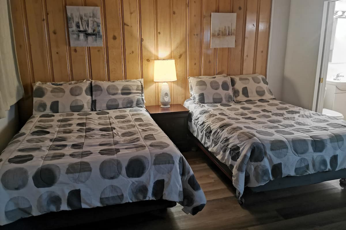 Una habitación con dos camas, paneles de madera en las paredes, una mesita de noche con lámpara y una ventana con vistas.