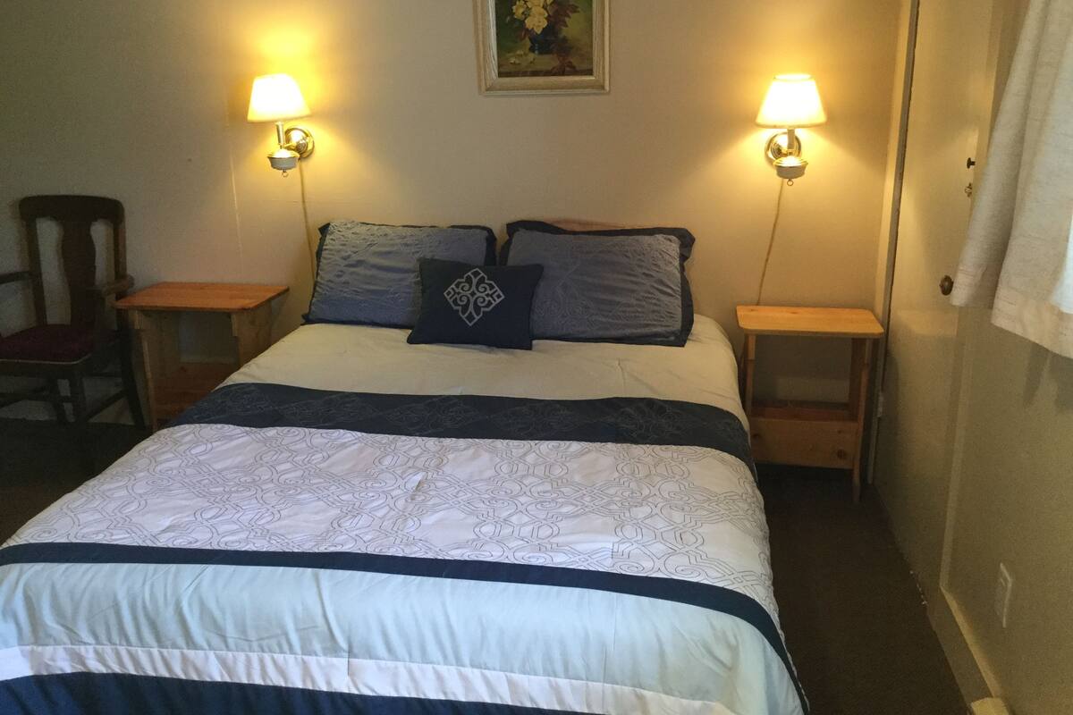 Una cama bien hecha con ropa de cama estampada, dos lámparas de noche, una silla y un cuadro en la pared.