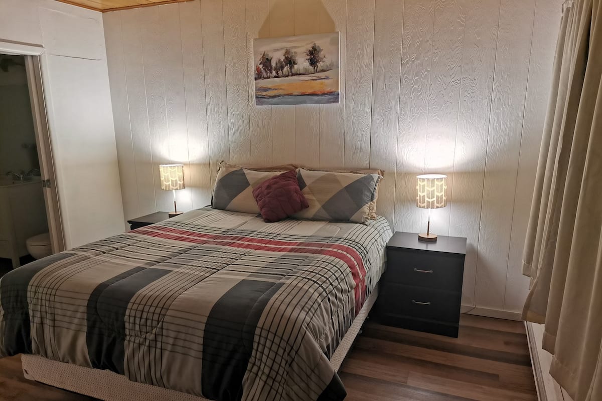 Un dormitorio con cama, dos mesitas de noche, un cuadro en la pared y una ventana con cortinas.