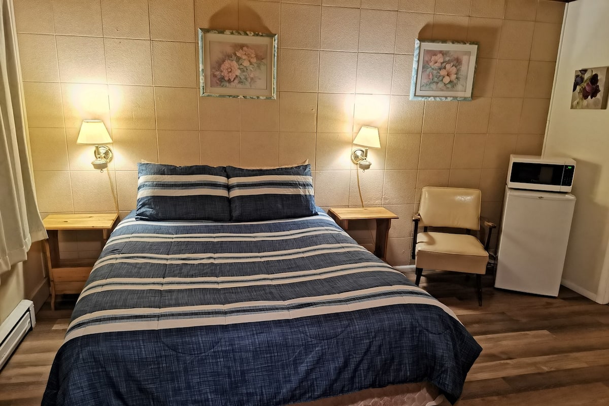 Un dormitorio con una cama, dos mesitas de noche, una silla, microondas y dos lámparas de pared.