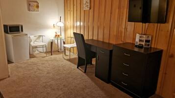 A small, cozy room with a wooden paneled wall, a flat-screen TV mounted above a dark wooden desk, a white refrigerator, a small table with a lamp, and two white chairs.
